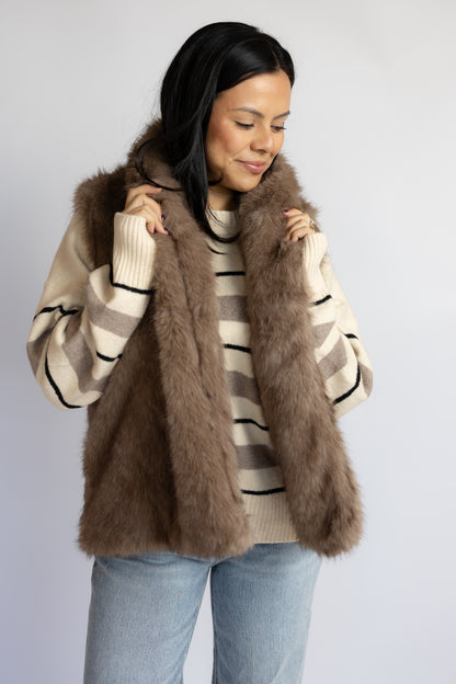 A woman wearing a brown faux fur vest over a cream and brown striped sweater, paired with light blue jeans, photographed against a plain light gray studio background.