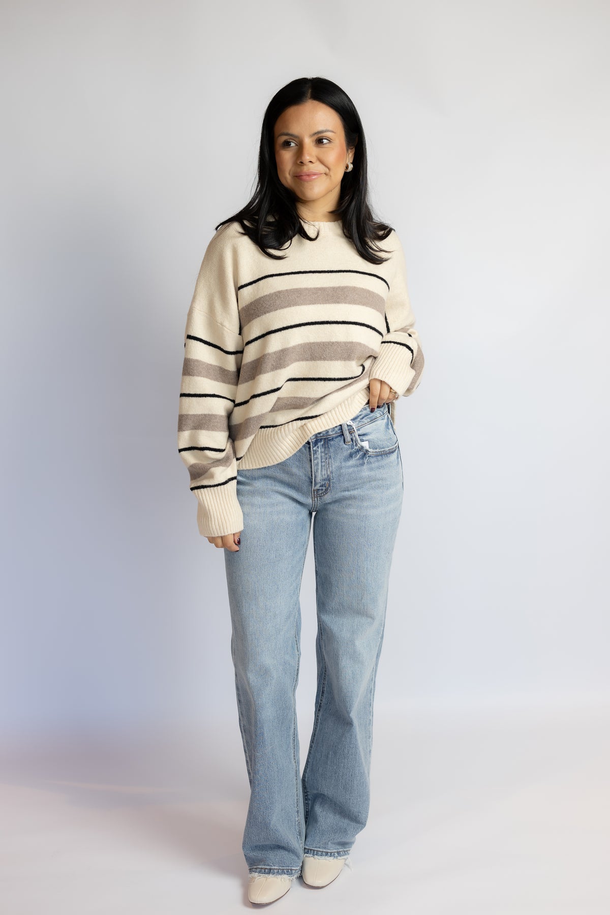 A woman modeling a cream and taupe striped knit sweater with light-wash blue jeans, standing against a light gray studio background.