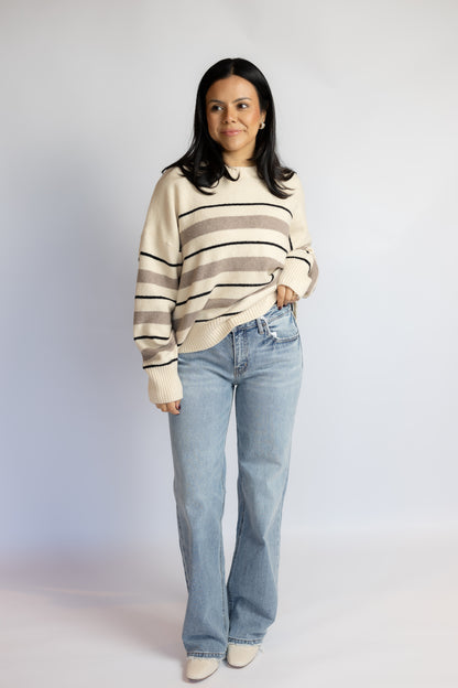 A woman modeling a cream and taupe striped knit sweater with light-wash blue jeans, standing against a light gray studio background.