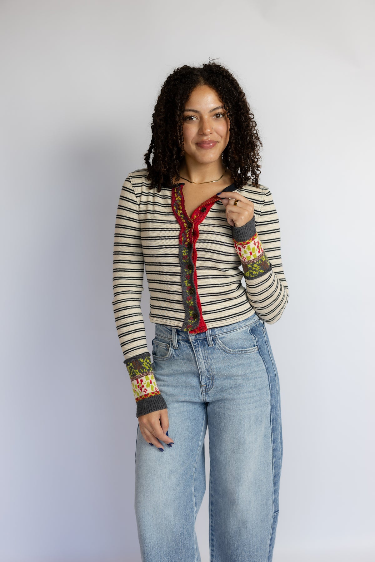 A woman with curly dark hair stands against a light gray studio background wearing a long-sleeve striped knit top with colorful patterned cuffs and a red button placket, paired with light blue jeans.