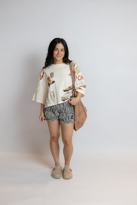 Woman wearing a white graphic t-shirt, patterned shorts, and beige shoes on a plain background