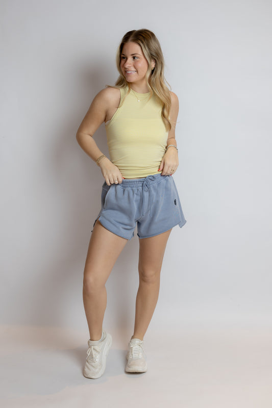 Woman wearing a yellow sleeveless top and blue shorts against a plain background