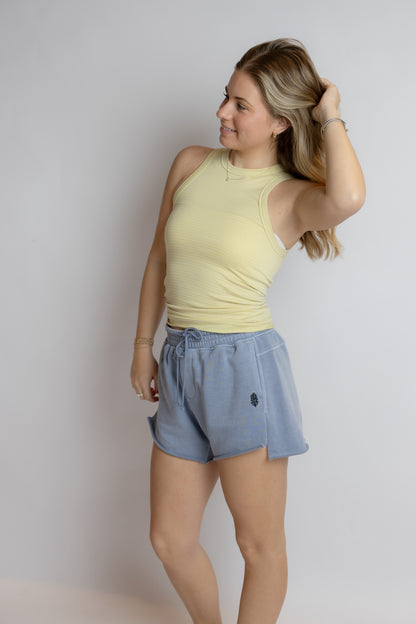 Woman wearing a yellow sleeveless top and blue shorts against a plain background