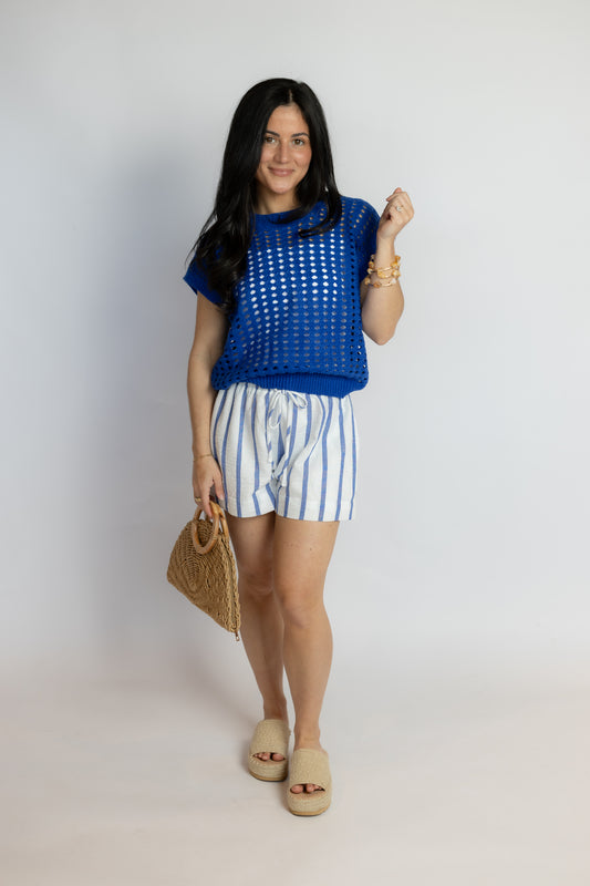 Woman wearing a blue top and striped shorts, holding a straw bag on a white background