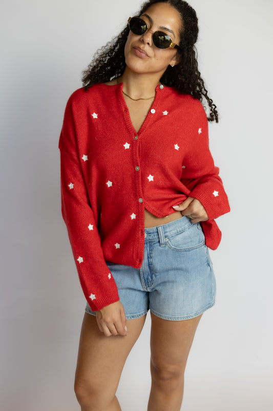 Woman wearing a red cardigan with white star patterns and denim shorts on a plain background