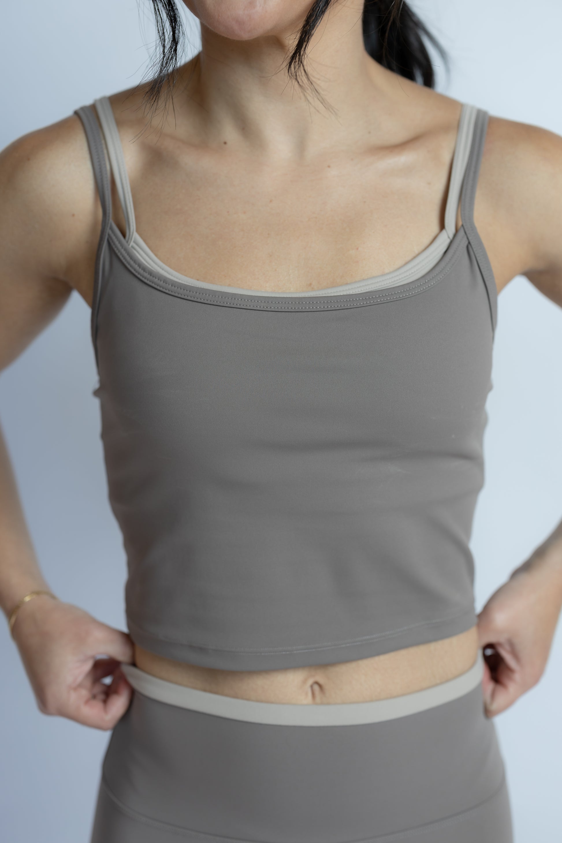 A woman wearing a taupe gray ribbed camisole tank top and matching high-waisted leggings, shown from the neck down against a light gray background.