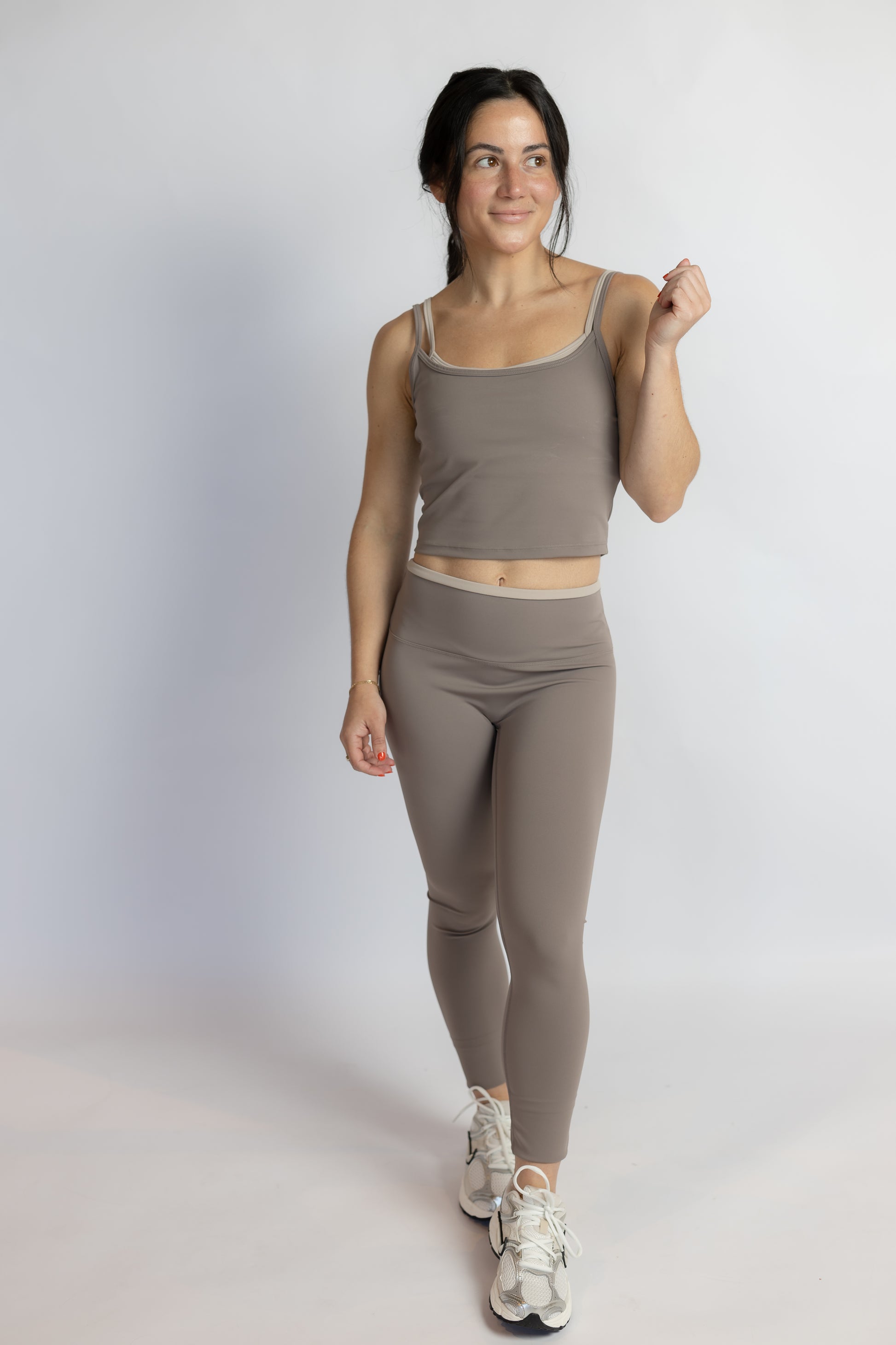A woman modeling a matching activewear set consisting of a taupe-brown tank top and high-waisted leggings, paired with white sneakers, against a light gray studio background.