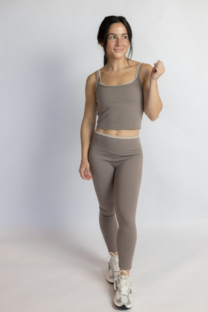 A woman modeling a matching activewear set consisting of a taupe-brown tank top and high-waisted leggings, paired with white sneakers, against a light gray studio background.
