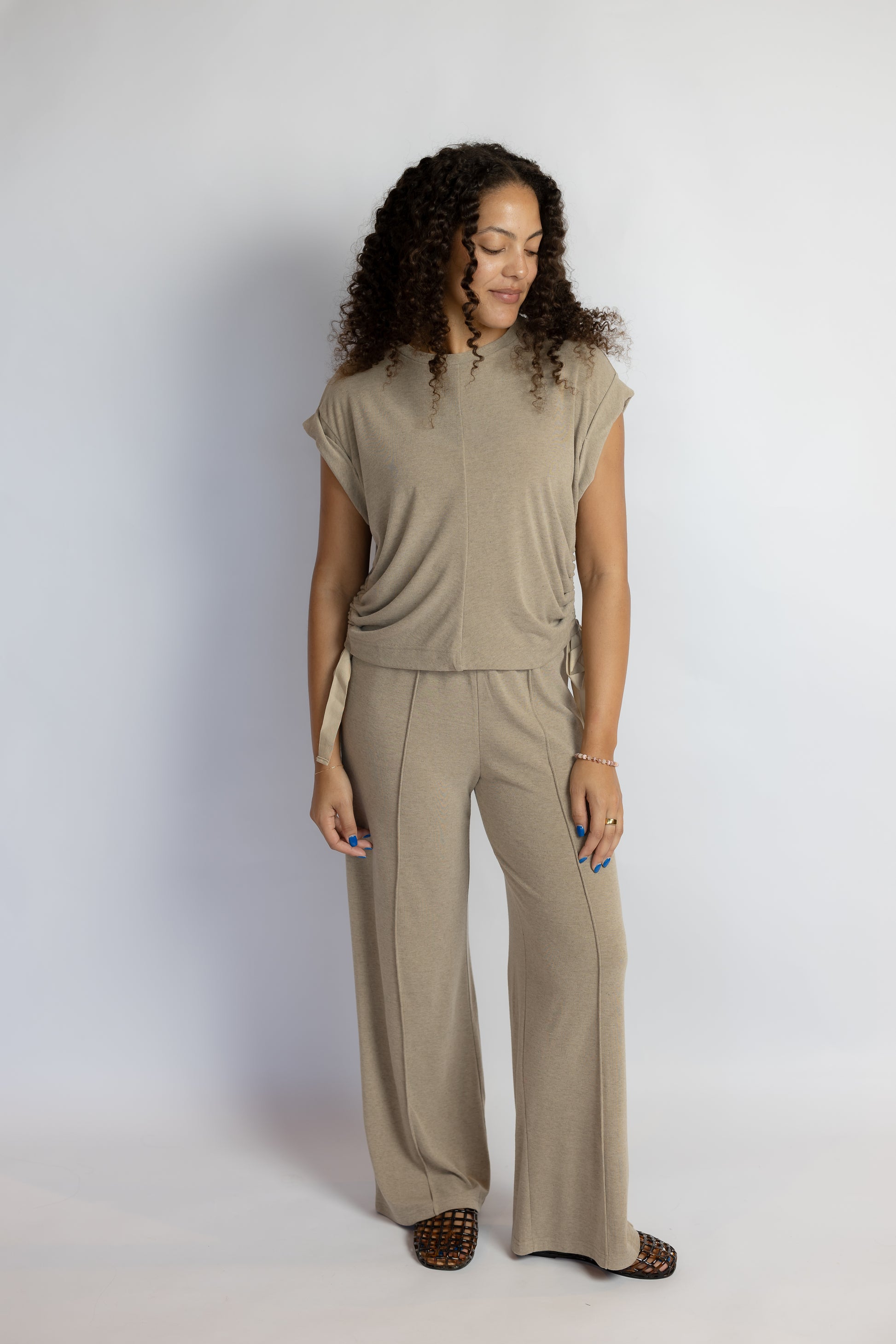 A woman modeling a matching beige two-piece lounge set consisting of a short-sleeve top and wide-leg pants, photographed against a light gray studio background.