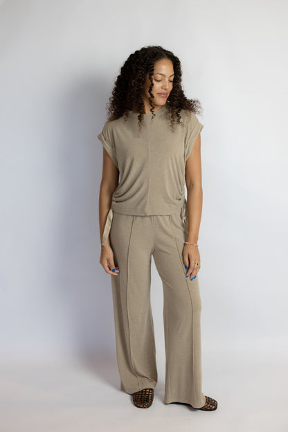 A woman modeling a matching beige two-piece lounge set consisting of a short-sleeve top and wide-leg pants, photographed against a light gray studio background.