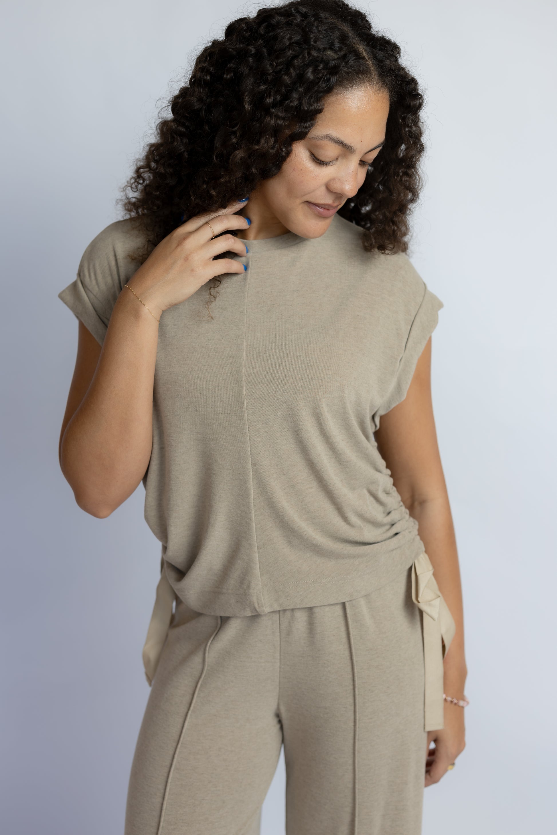 A woman with curly dark hair wearing a light taupe knit top and matching knit pants, photographed against a plain light gray studio background.