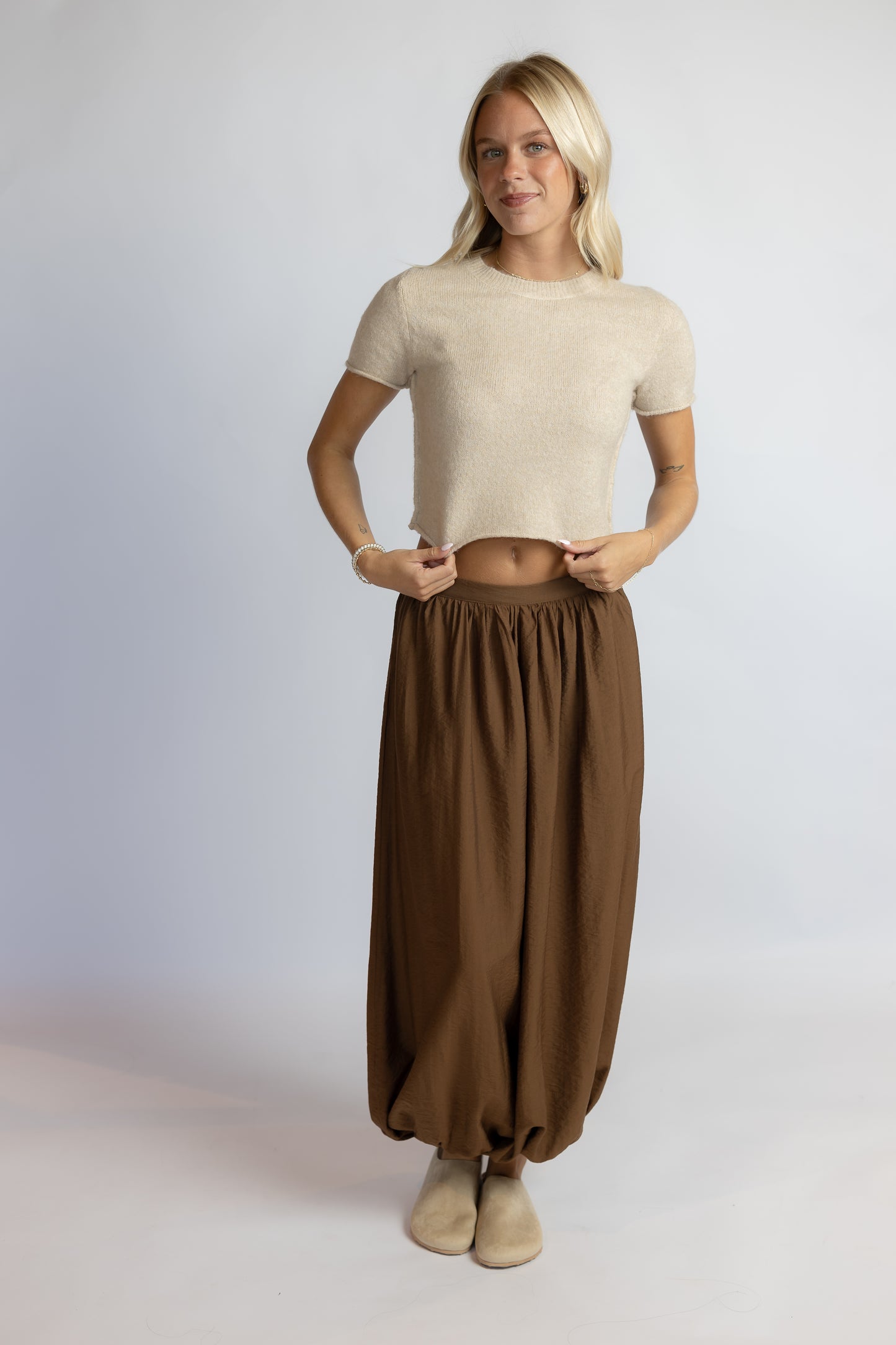 A woman modeling a beige short-sleeve knit crop top and brown harem-style pants against a light gray studio background.