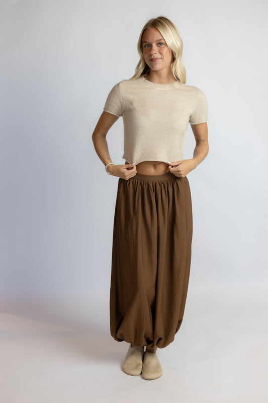 A woman modeling a beige short-sleeve knit crop top and brown harem-style pants against a light gray studio background.