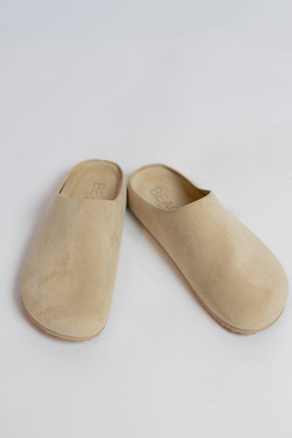 A pair of beige suede mule slip-on shoes with a minimalist design, photographed on a clean white background.