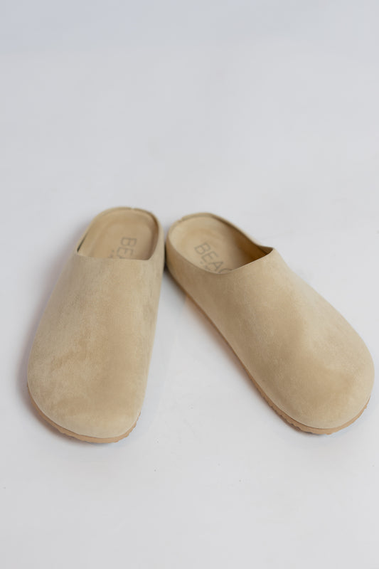 A pair of beige suede mule slip-on shoes with a minimalist design, photographed on a clean white background.