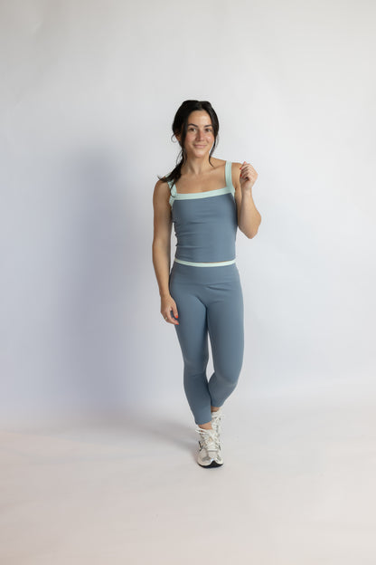 A woman modeling a matching slate blue activewear set consisting of a square-neck tank top and high-waisted leggings, with light mint green trim, set against a plain light gray studio background.