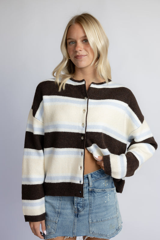 A woman modeling a brown, cream, and light blue striped knit cardigan with a button-up front, paired with a denim mini skirt, against a plain light gray studio background.