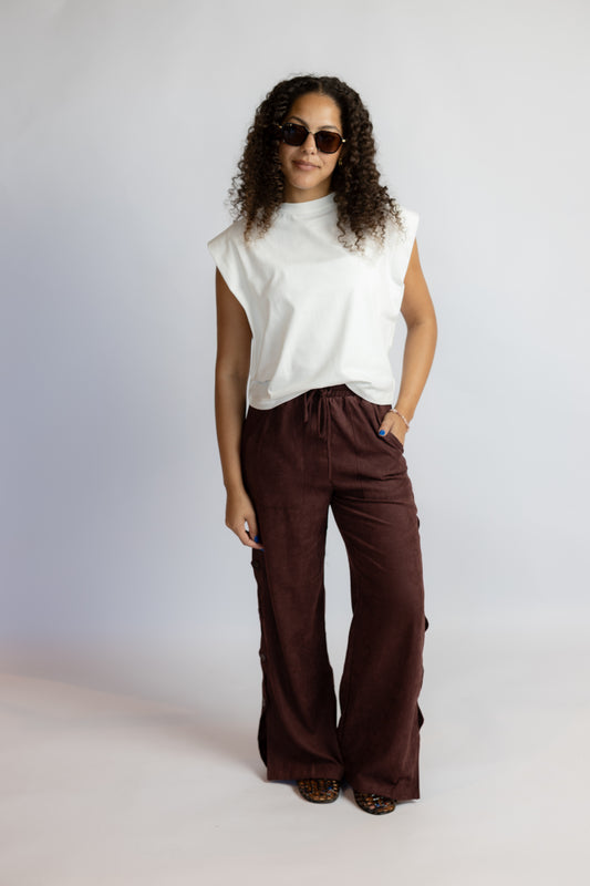 A woman modeling a white sleeveless mock-neck top and dark brown wide-leg pants against a light gray studio background.