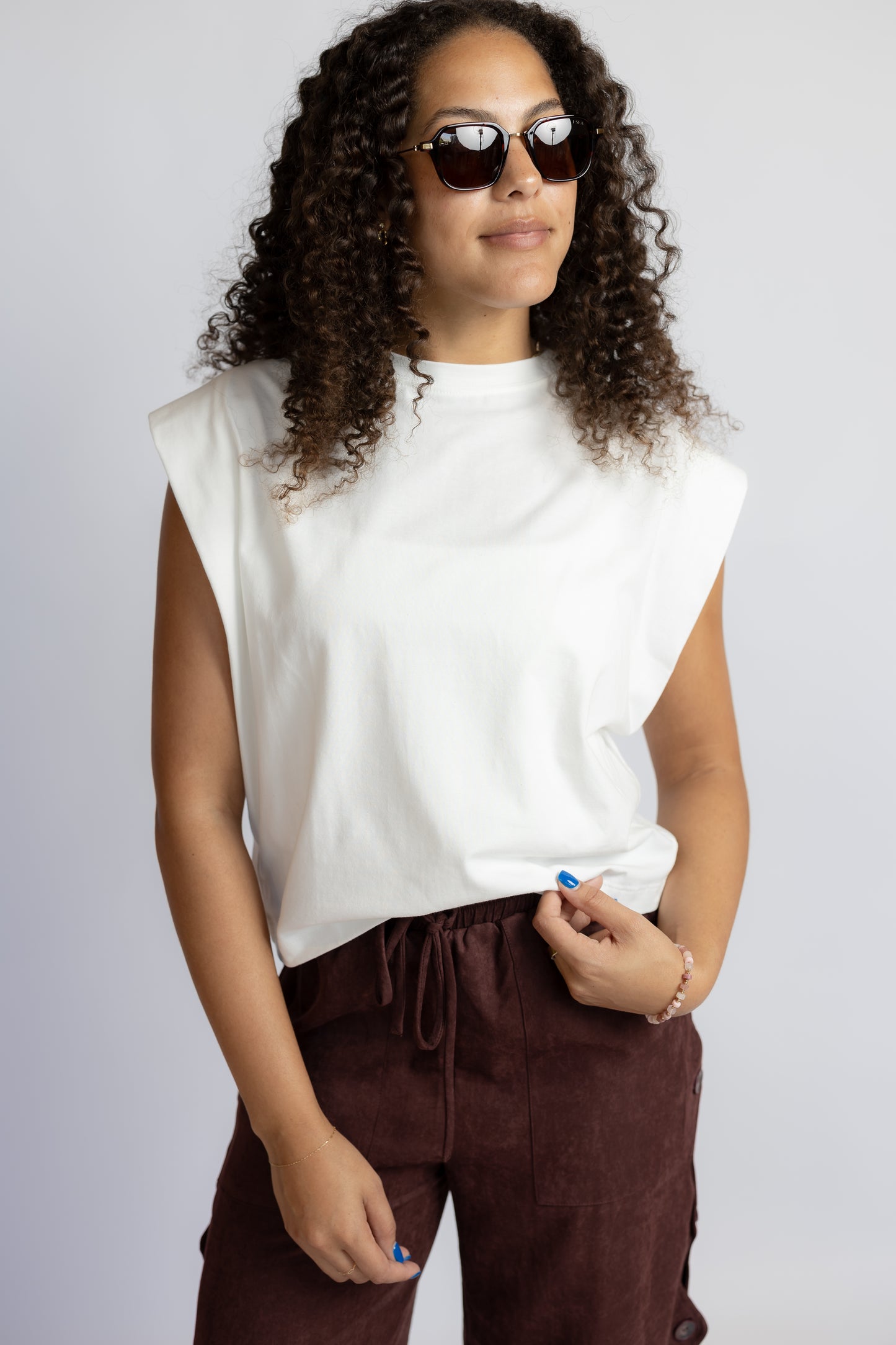 A woman with curly hair wearing a white sleeveless crop top and dark brown pants, posing against a light gray studio background.