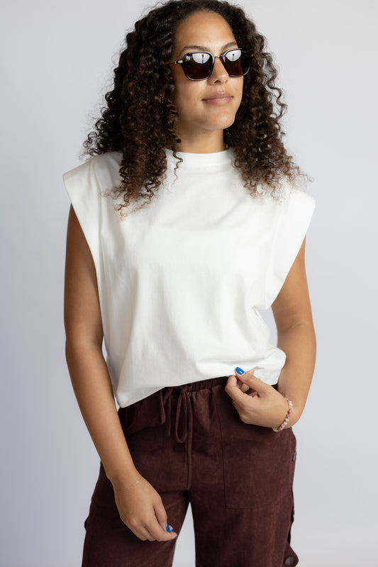 A woman with curly hair wearing a white sleeveless crop top and dark brown pants, posing against a light gray studio background.