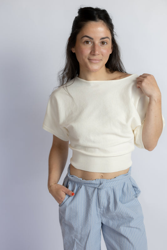 A woman modeling a cream-colored short-sleeve crop top with a wide boat neckline, paired with light blue and white striped pants, against a plain light gray background.