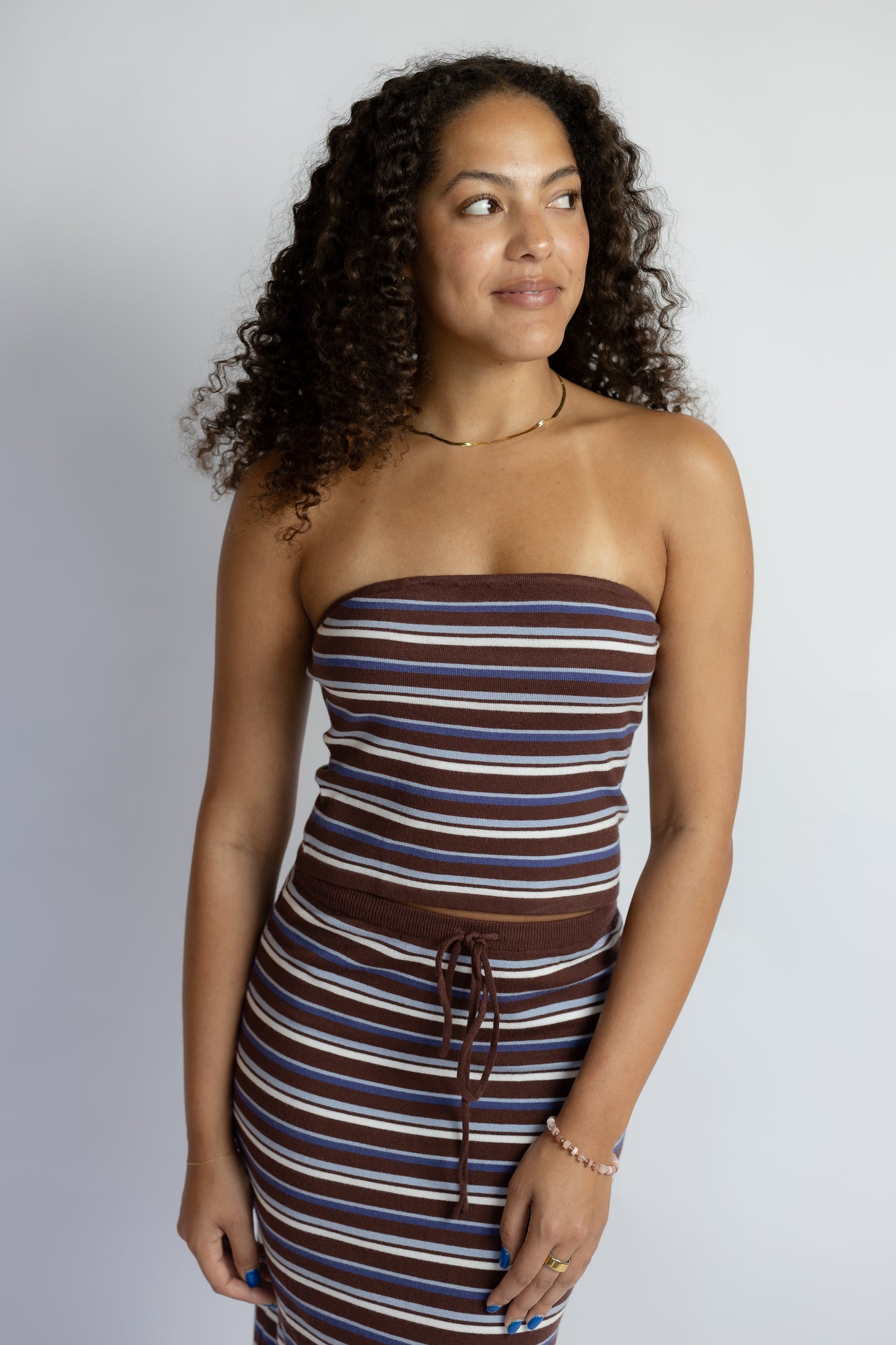 A woman with curly dark hair wearing a brown, white, and blue striped strapless tube dress with a drawstring waist, posing against a light gray studio background.