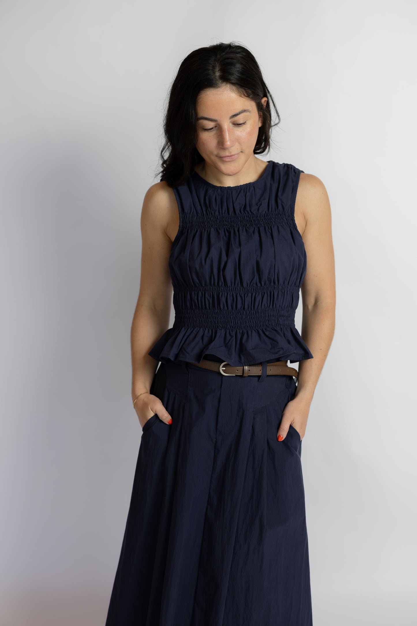 A woman wearing a sleeveless navy blue smocked peplum top and matching navy blue wide-leg pants, styled with a brown belt, standing against a light gray studio background.