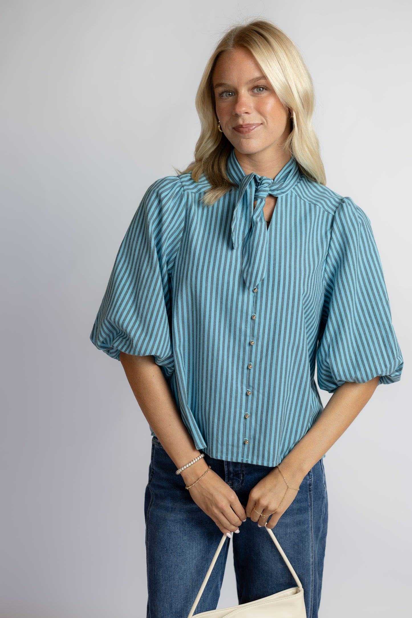 A woman modeling a blue and white vertically striped blouse with puff sleeves, a tie-neck detail, and front buttons, paired with blue jeans against a light gray studio background.