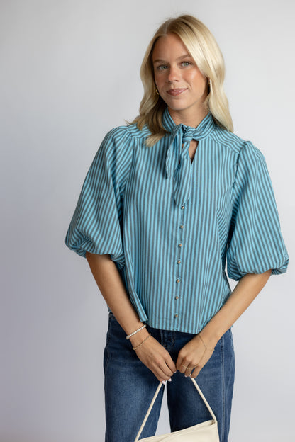 A woman modeling a blue and white vertically striped blouse with puff sleeves, a tie-neck detail, and front buttons, paired with blue jeans against a light gray studio background.