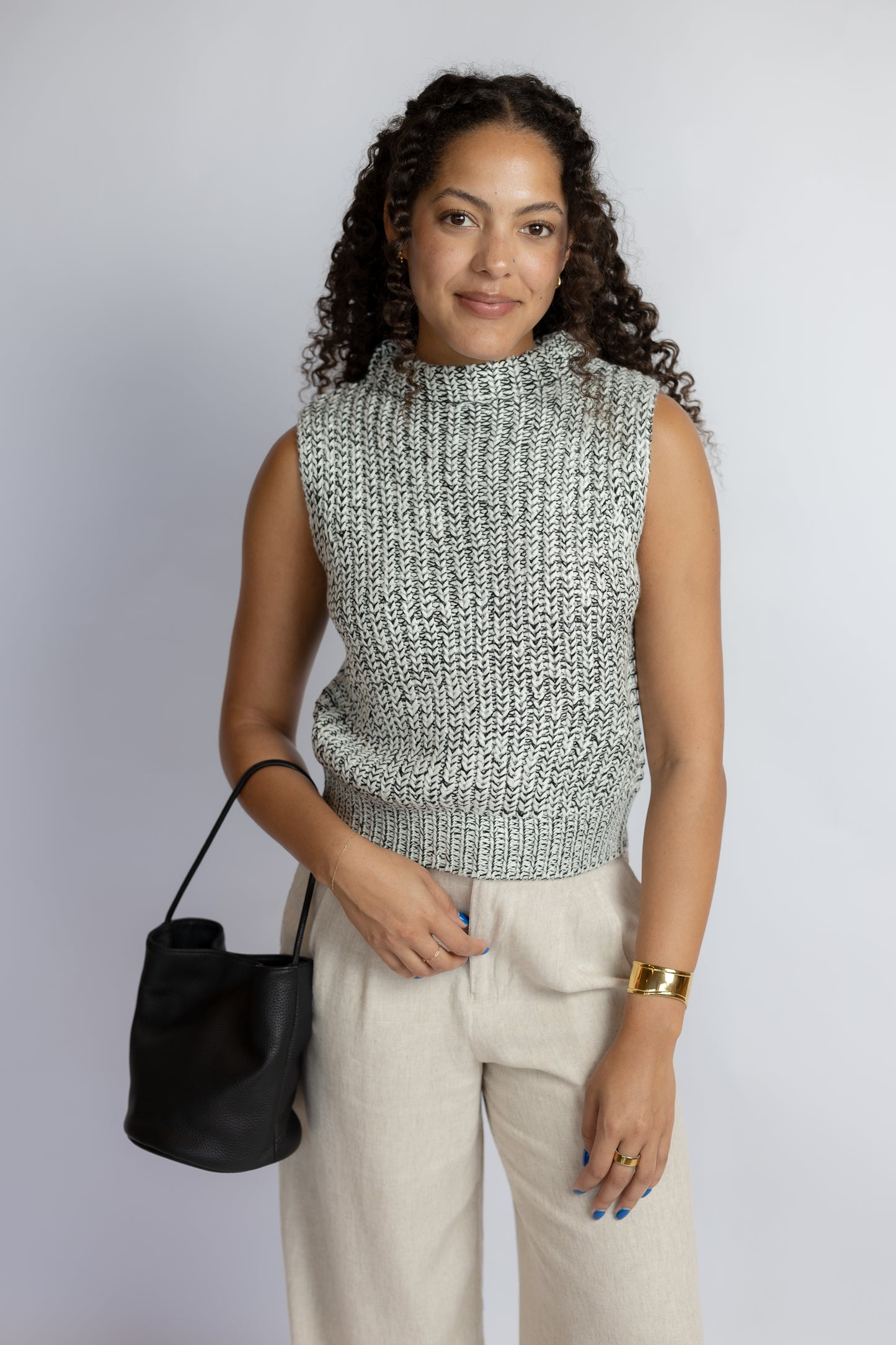 A woman with curly dark hair models a sleeveless, high-neck, textured knit sweater vest in a black and white marled pattern, paired with light beige trousers and a black leather bucket bag, against a plain light gray studio background.