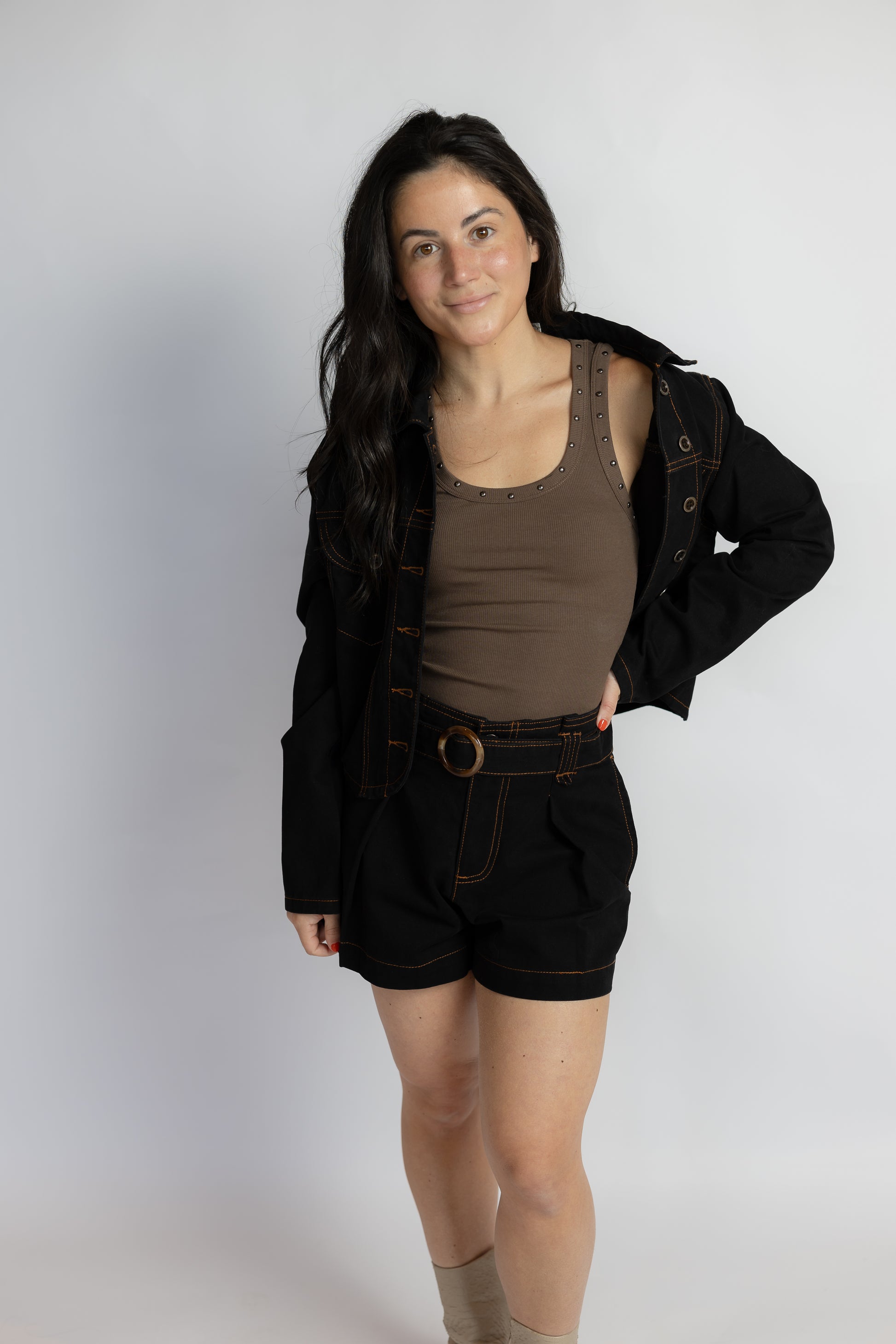 A woman modeling a black denim jacket, brown ribbed tank top, and black denim shorts with a belt, standing against a light gray studio background.