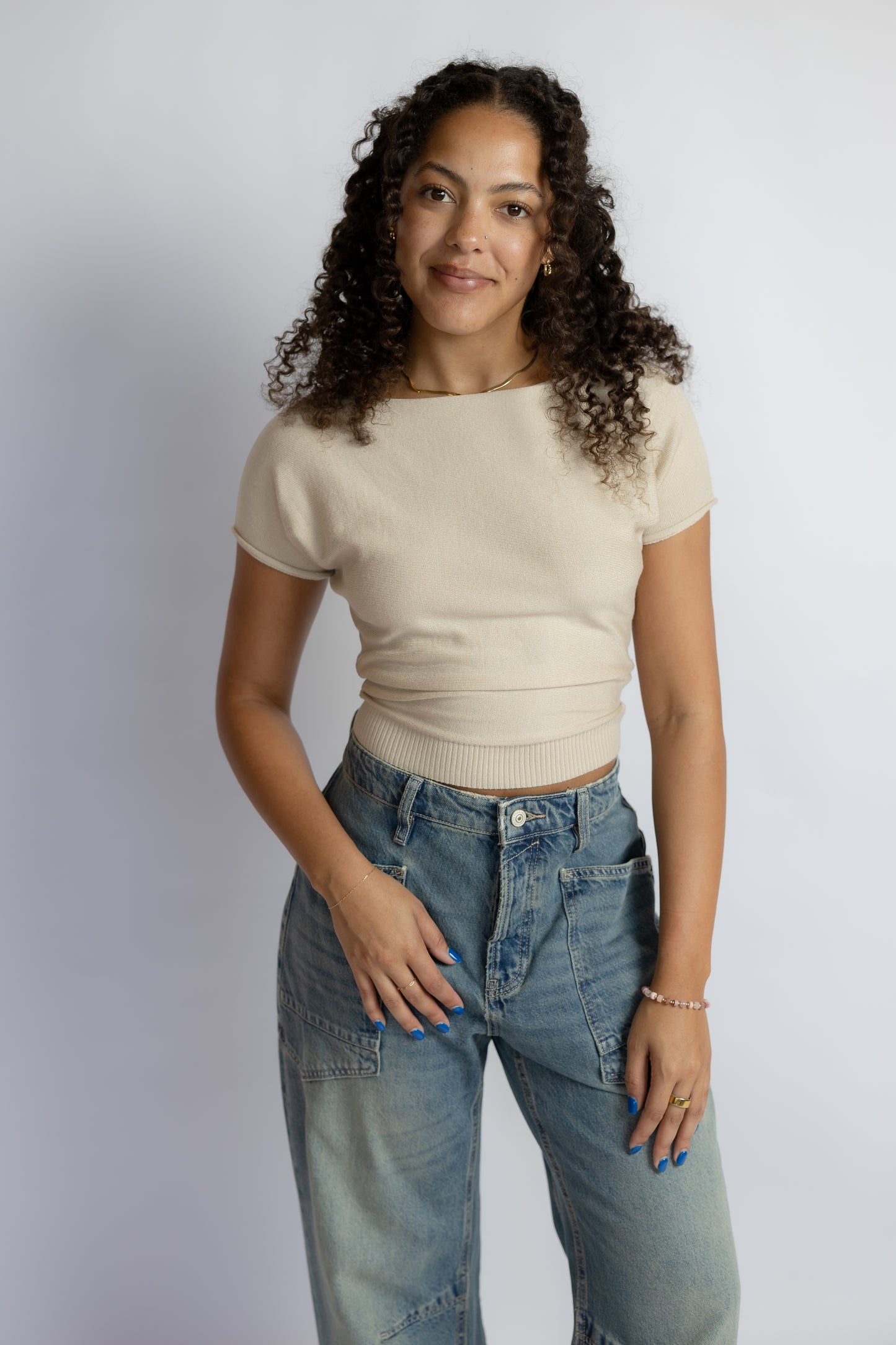 A woman with curly dark hair wearing a beige short-sleeve knit top and light blue jeans, standing against a light gray studio background.