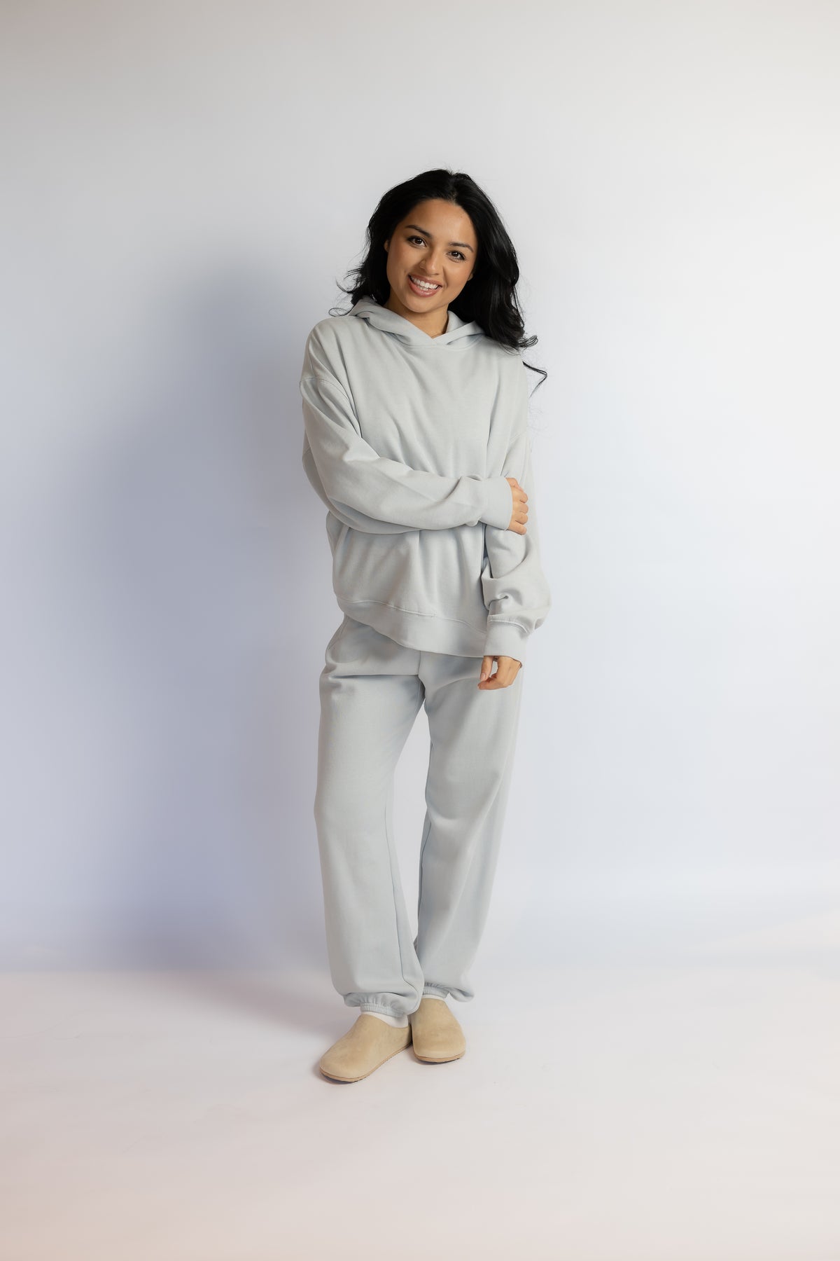 A woman modeling a light gray two-piece loungewear set consisting of a hooded sweatshirt and matching sweatpants, paired with beige slip-on shoes, against a plain light gray studio background.