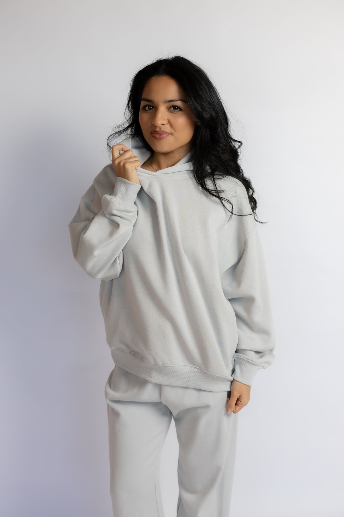 A woman modeling a light gray matching lounge set consisting of a sweatshirt and sweatpants, standing against a plain light gray studio background.