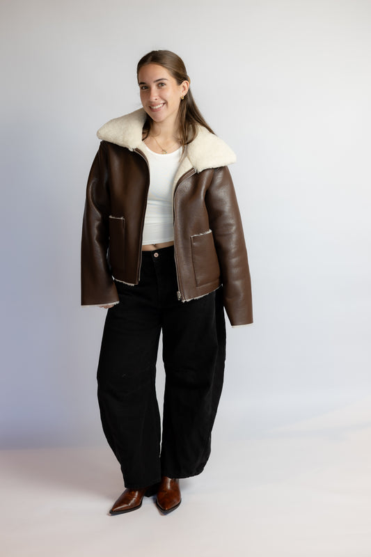 A woman modeling a brown leather aviator jacket with a thick cream shearling-style collar, worn over a white top and black wide-leg pants, against a light gray studio background.