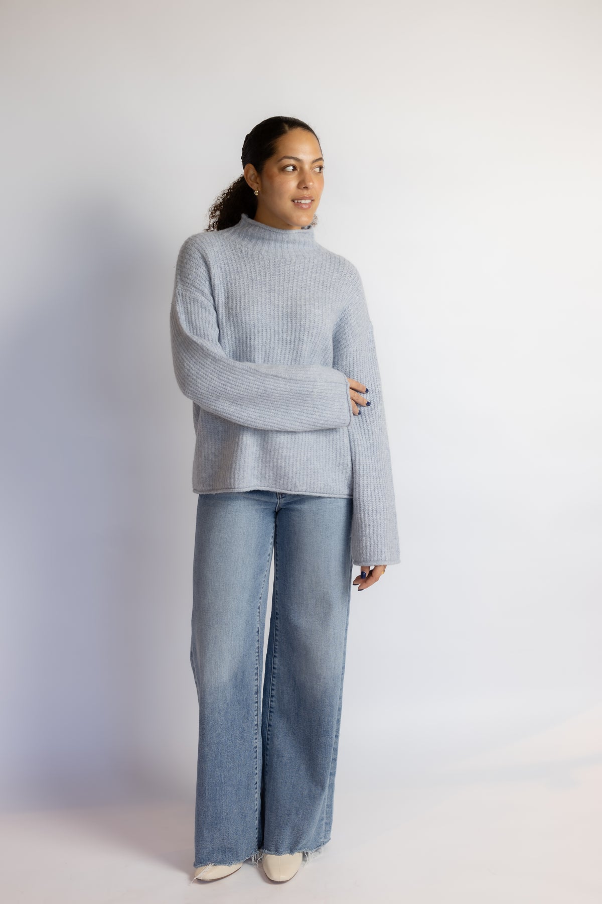 A woman modeling a light blue ribbed knit mock-neck sweater and wide-leg blue jeans against a plain white studio background.