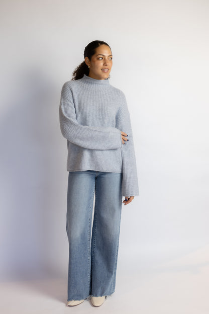 A woman modeling a light blue ribbed knit mock-neck sweater and wide-leg blue jeans against a plain white studio background.