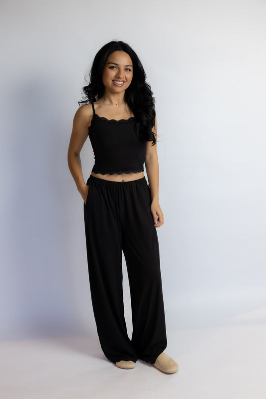 A woman modeling a black two-piece loungewear set consisting of a cropped camisole top with scalloped edges and matching wide-leg pants, paired with beige slip-on shoes, against a light gray studio background.