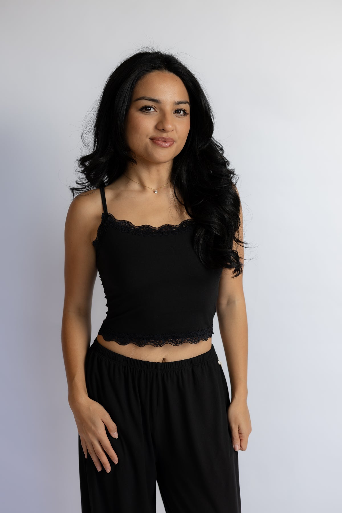 A woman with long, wavy black hair is wearing a black camisole top with a scalloped lace trim and black pants, standing against a plain light gray background.