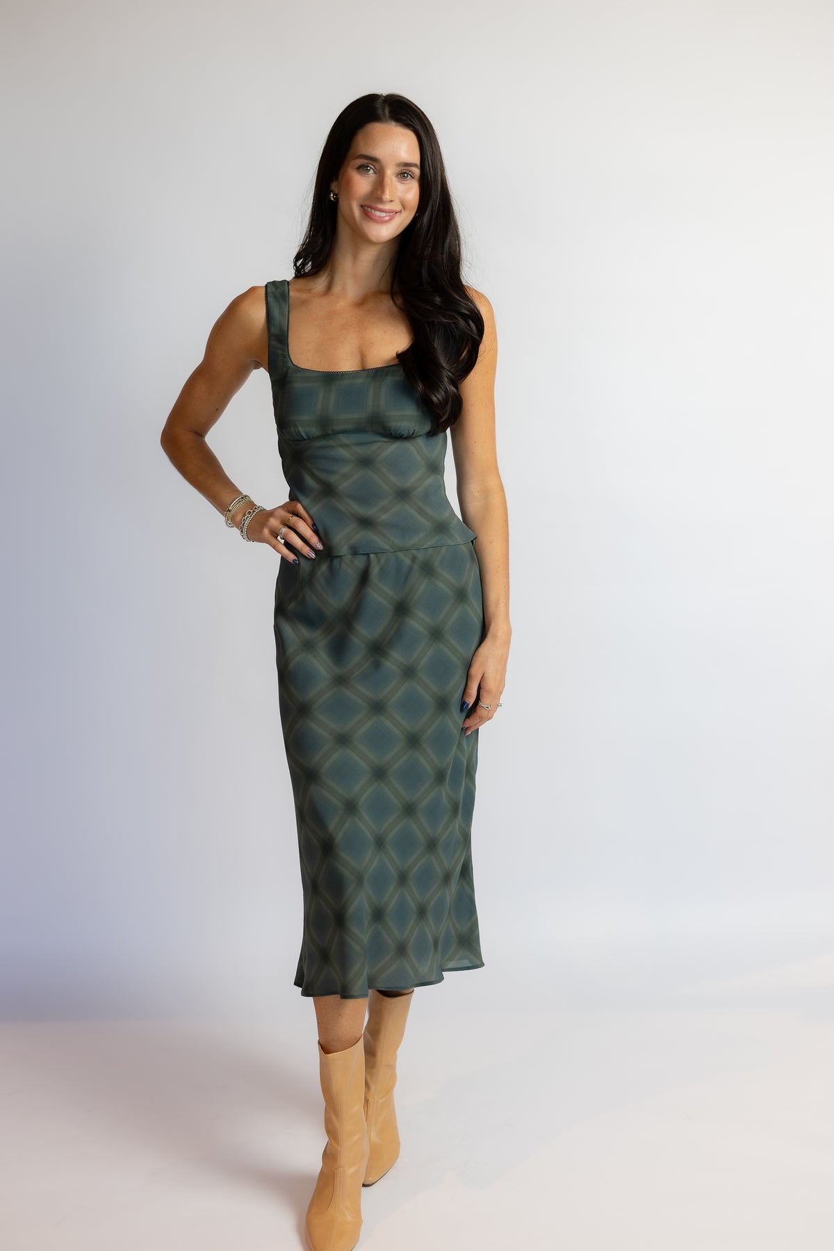 A woman modeling a sleeveless midi dress with a green and blue geometric pattern, paired with tan ankle boots, against a plain light gray studio background.