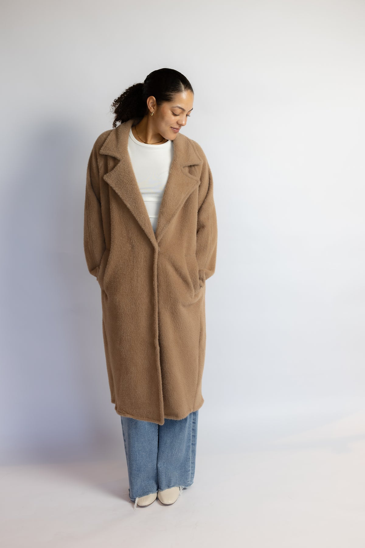 A woman wearing a long camel-colored wool coat over a white top and wide-leg blue jeans, standing against a plain light gray studio background.