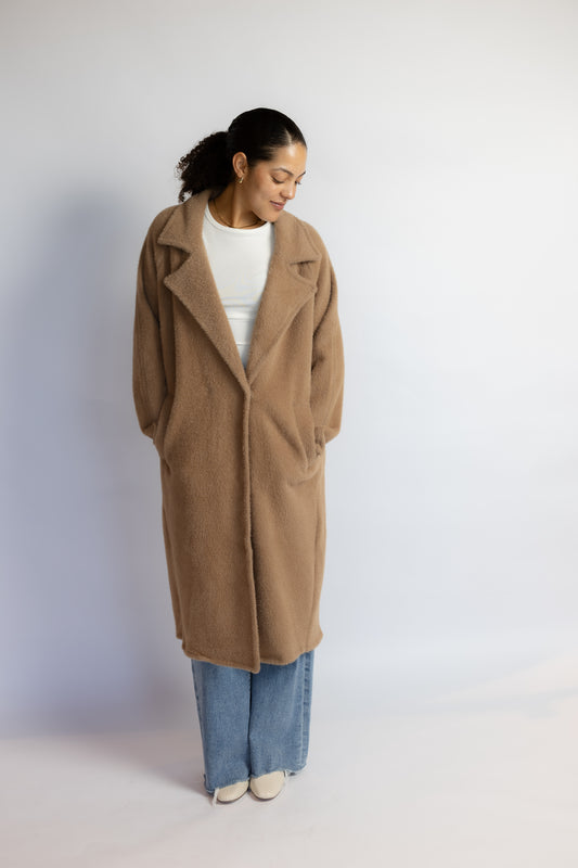 A woman wearing a long camel-colored wool coat over a white top and wide-leg blue jeans, standing against a plain light gray studio background.
