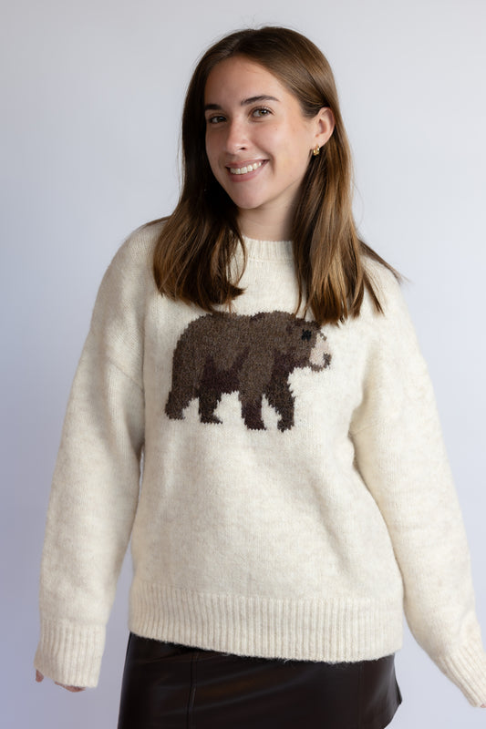 A woman wearing a cream-colored knit sweater with a brown bear motif across the chest, paired with black bottoms, set against a plain light gray background.
