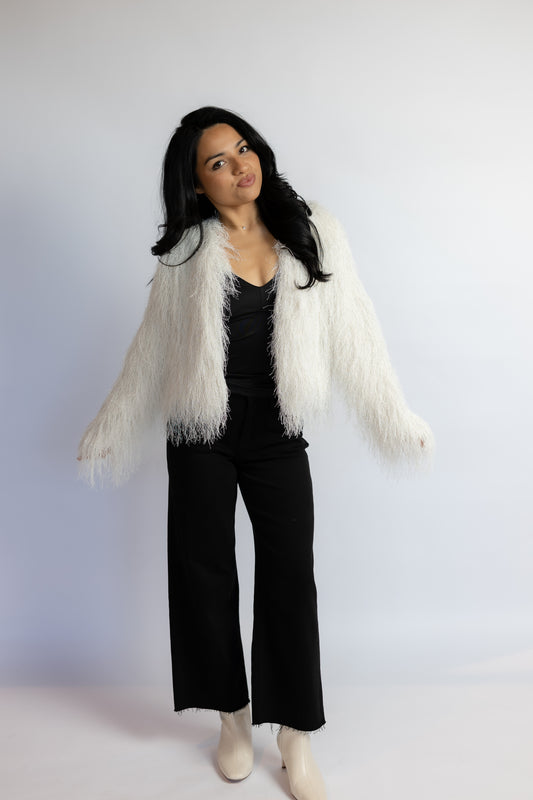 A woman modeling a white shaggy faux-fur cropped jacket over a black top and black wide-leg pants, standing against a plain light gray studio background.