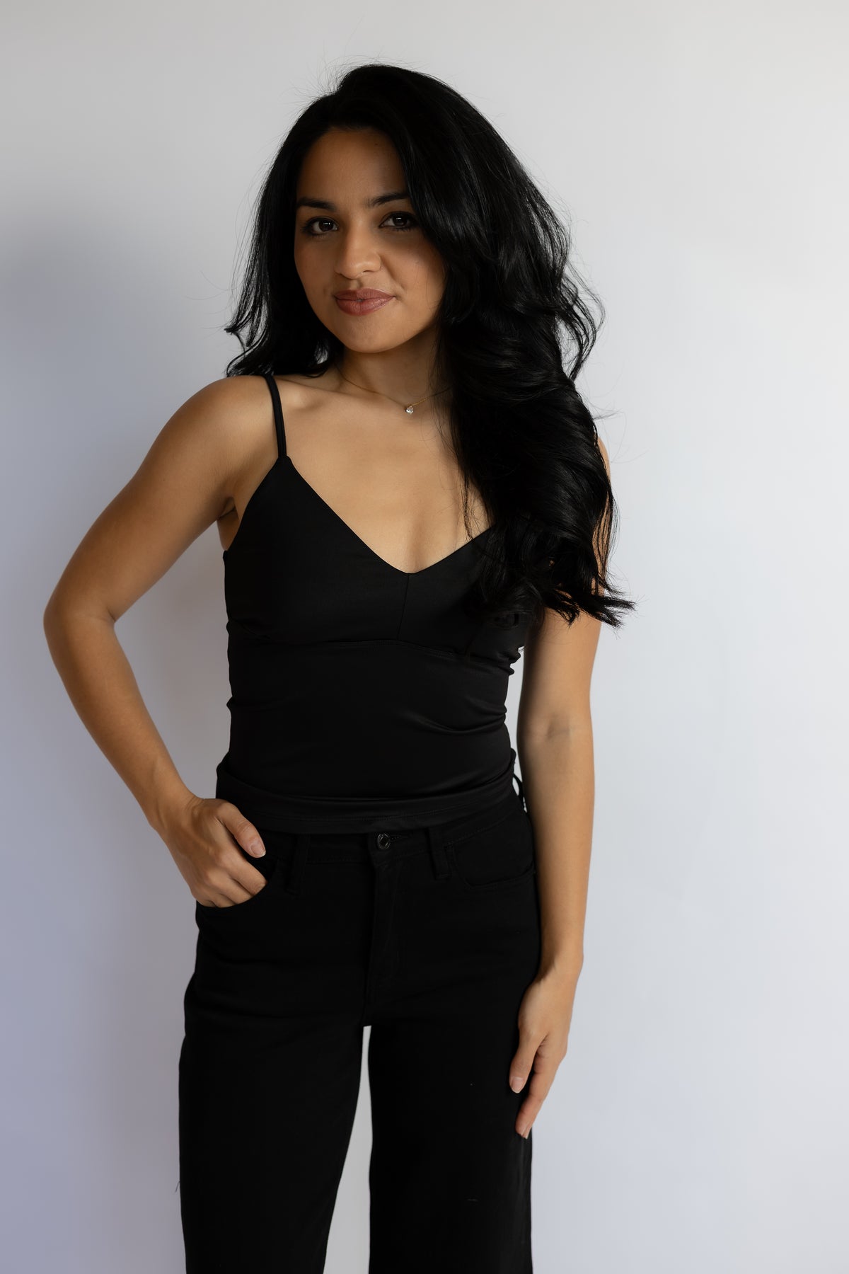 A woman with long, wavy black hair is wearing a black spaghetti-strap camisole top and black pants, standing against a plain light gray wall.