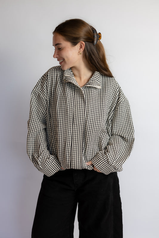 A woman wearing a black and white gingham long-sleeve button-up shirt and black pants, standing against a plain light gray wall.