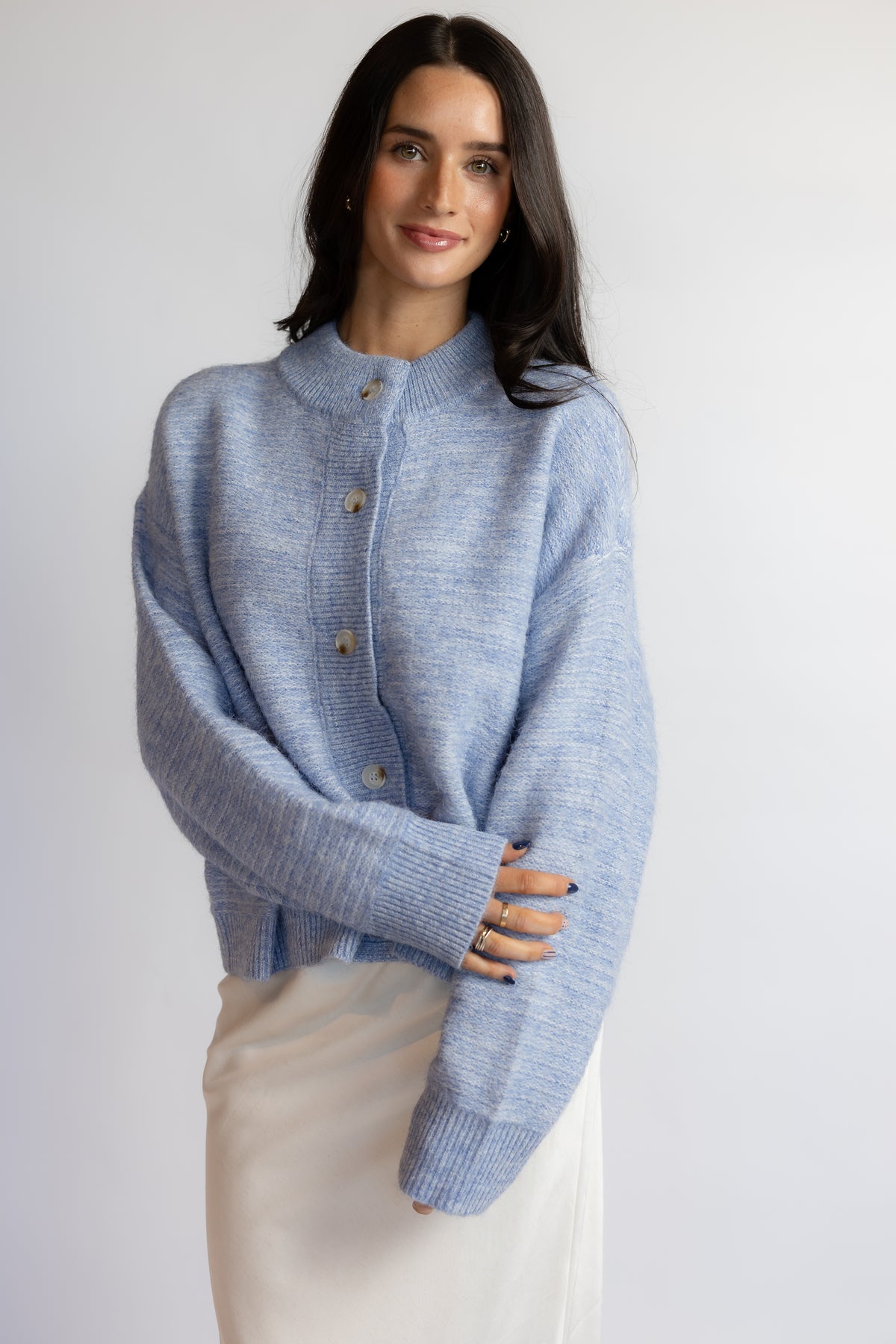 A woman wearing a light blue knit cardigan sweater with a mock neck and brown buttons, paired with a cream-colored skirt, photographed against a plain light gray background.