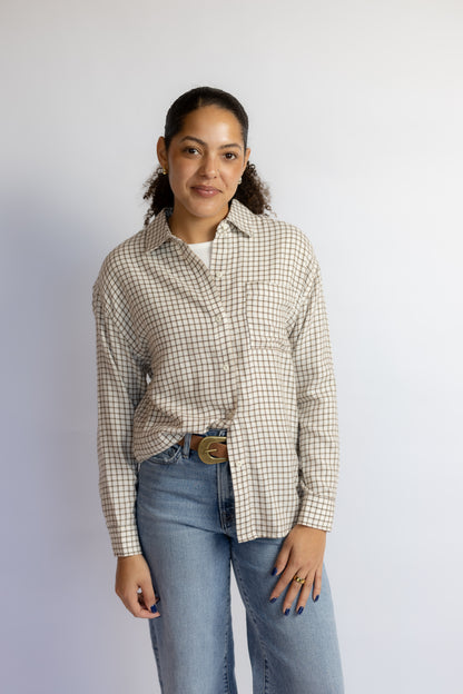 A woman modeling a beige and brown gingham long-sleeve button-up shirt with blue jeans, standing against a plain light gray studio background.