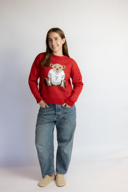 A woman modeling a red crewneck sweater featuring a teddy bear graphic wearing a white jersey with the number 7, paired with blue jeans and beige slip-on shoes, against a plain light gray studio background.