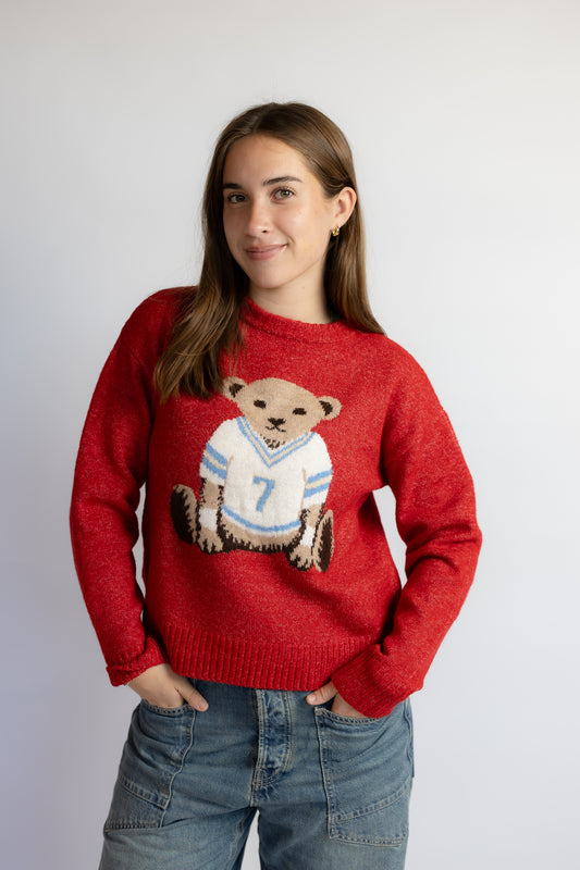 A woman wearing a red crewneck sweater with a knitted graphic of a teddy bear in a football jersey with the number 7, paired with blue jeans, against a plain light gray background.